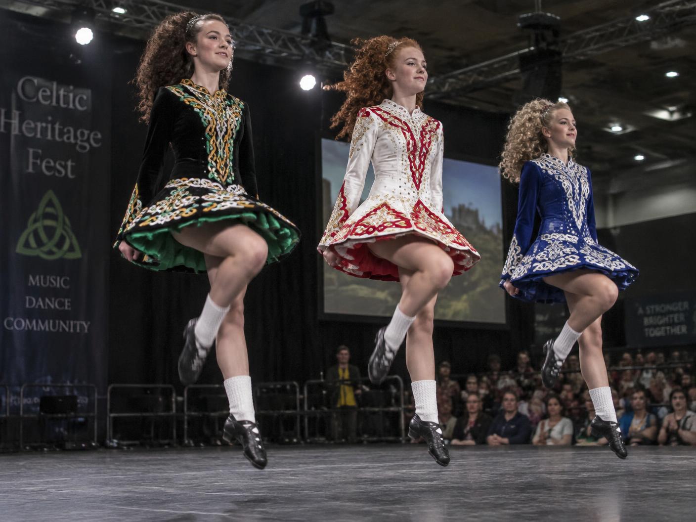 Three Irish step dancers in embroidered dresses caught mid-leap on an indoor stage before a seated audience