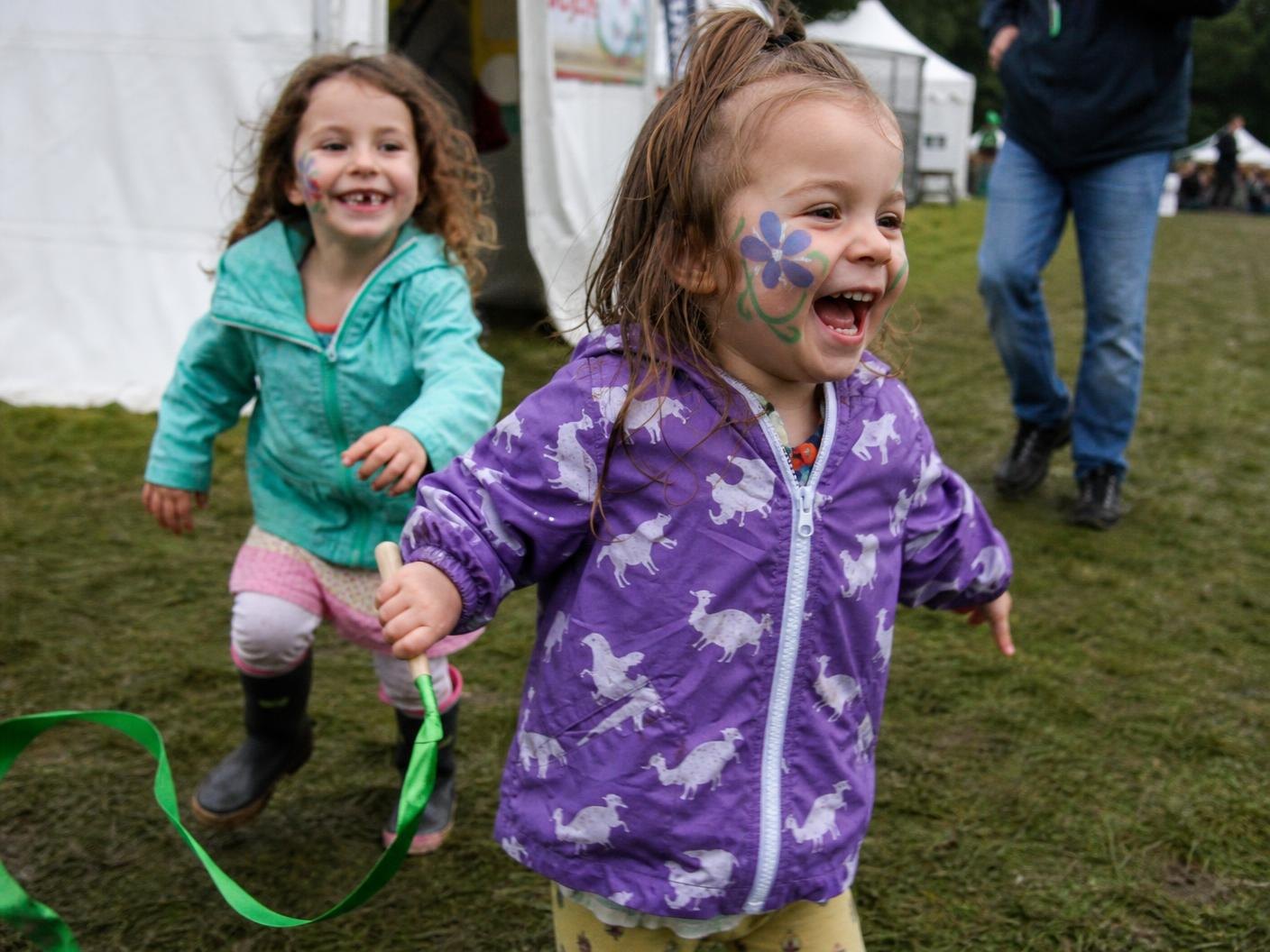 Two small children with painted faces running and laughing with a green ribbon streamer on festival grass