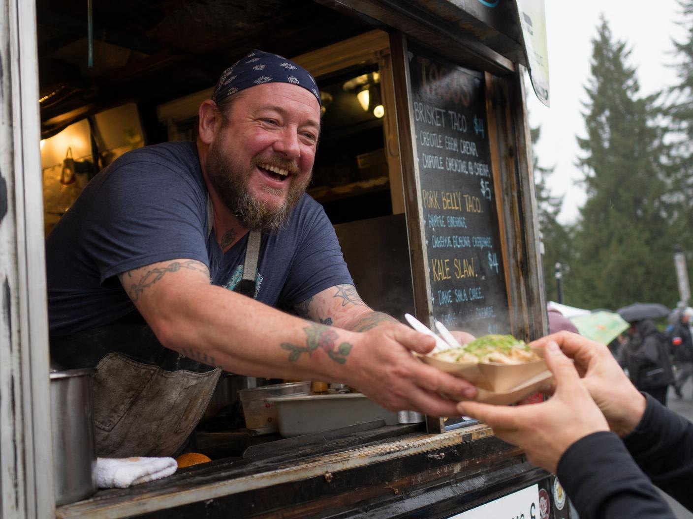 A laughing cook leaning out of a food stall window to hand a tray of food to a visitor
