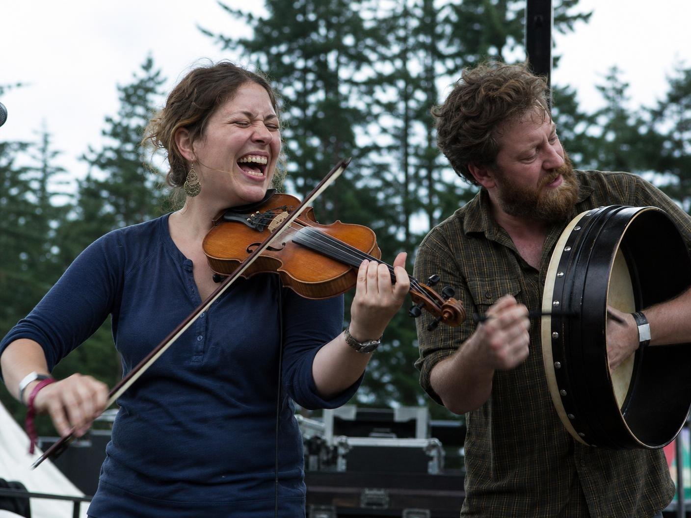 A fiddler laughing mid-tune beside a bodhrán player on an outdoor stage backed by Douglas fir